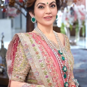 May include: A woman wearing a pink and gold sari with intricate embroidery and embellishments. She is adorned with diamond necklaces and emerald earrings and bracelets. A green bindi is on her forehead.