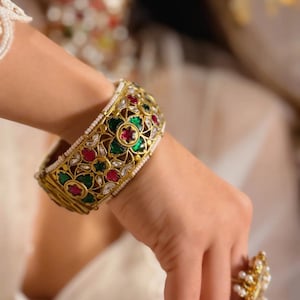 May include: A wide gold bangle bracelet featuring intricate floral cutouts, inlaid with vibrant green and red stones, and accented with small white pearls along the edges. A matching gold ring with white pearls is also visible on the finger.