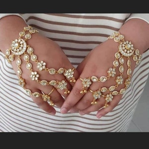 May include: A pair of gold-toned, ornate, hand jewelry sets with white stones. Each set features a circular design with a dangling chain and multiple flower-shaped charms.