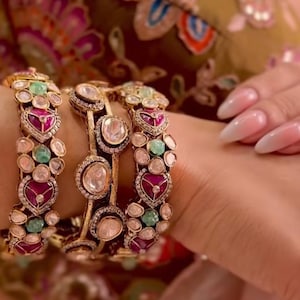 Op de afbeelding: Drie goudkleurige armbanden met een herhalend patroon van hartvormige magentakleurige stenen, ronde doorschijnende witte stenen en kleine ronde groene stenen. De metalen zettingen zijn afgewerkt met kleine witte accenten langs de randen.