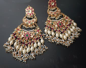 Puede incluir: Pendientes colgantes dorados ornamentados. Cada pendiente presenta un diseño en forma de media luna adornado con piedras preciosas rojas, verdes y blancas, y perlas de imitación colgantes. Los pendientes tienen un diseño floral en la parte superior.