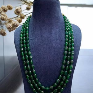 Puede incluir: Collar de varias hebras con cuentas verde oscuro, exhibido en un maniquí azul. Las cuentas pulidas están dispuestas en capas. El fondo presenta una pared neutra y elementos decorativos. El collar es una pieza llamativa.
