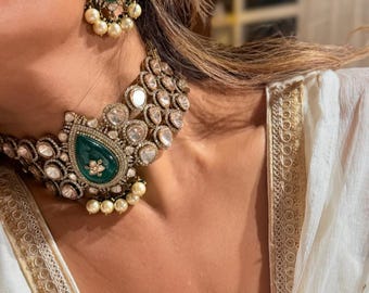 May include: A luxurious jewelry set featuring a choker necklace and matching earrings. The necklace and earrings have a silver-tone metal setting with clear and emerald-green gemstones and small pearl accents. The jewelry is worn with a white garment.