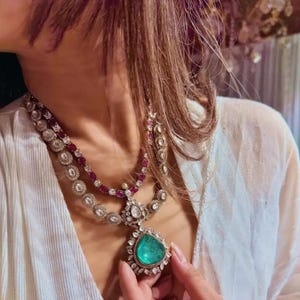 May include: A layered silver necklace with a large emerald green gemstone pendant. The necklace features a row of small red gemstones and a row of white gemstones.