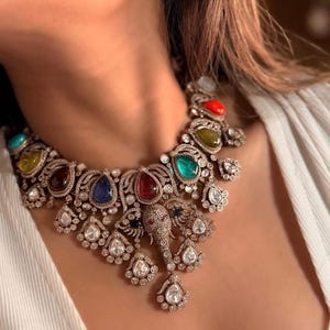 May include: A statement necklace featuring a central elephant motif crafted in a metallic finish with intricate detailing. The piece is adorned with multiple teardrop-shaped gemstones in red, blue, green, yellow, and orange, accented by small clear stones.