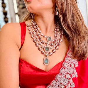 Pode incluir: Um colar de várias camadas com pedras de cor esmeralda e um par de brincos a condizer. O colar é cravejado com pequenas pedras transparentes e detalhes vermelhos. A mulher está vestindo um top vermelho e uma peça bordada em vermelho e branco.