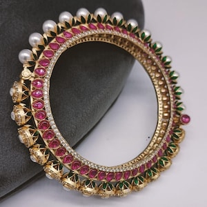 Może przedstawiać: Złota bransoletka obręczowa ze skomplikowanymi detalami. Bransoletka ma naprzemiennie rubinowe kamienie, przezroczyste kryształy i białe perły. Zielone i złote akcenty dopełniają design. Biżuteria jest umieszczona na szarym tle.