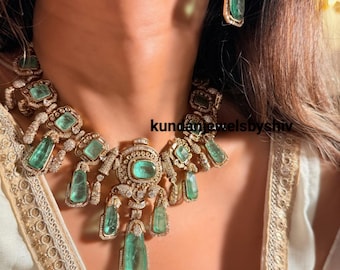 May include: A luxurious necklace and earring set featuring large, rectangular emeralds and small diamonds set in gold. The necklace has a central oval emerald and dangling emerald pendants. The earrings have a similar design. The text "kundanjewelsbyshiv" is visible.
