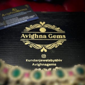 May include: A black box with a gold logo that says "Avighna Gems" and the text "Kundanjewelsbyshiv Avighnagems". The box is open and a gold necklace with green and pink stones is visible.