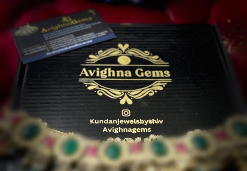 May include: A black box with a gold logo that says "Avighna Gems" and the text "Kundanjewelsbyshiv Avighnagems". The box is open and a gold necklace with green and pink stones is visible.
