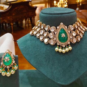 May include: A gold necklace with a large emerald green gemstone pendant surrounded by smaller diamonds. The necklace is on a green velvet display stand. A pair of matching earrings with emerald green gemstones and pearl accents are also shown.