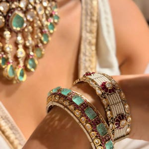 May include: Two ornate metal bangles featuring rectangular green stones, small red circular accents, and white seed bead rows. A matching heavy necklace with teardrop-shaped green stones and red accents is visible in the background, creating a set.
