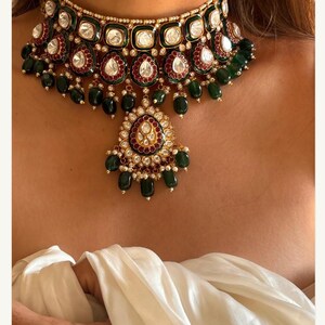 May include: A luxurious choker necklace featuring a gold-toned setting with emerald green and ruby red accents. The necklace is adorned with clear, square-cut stones and teardrop-shaped emerald green beads. A pendant with a similar design hangs from the center.