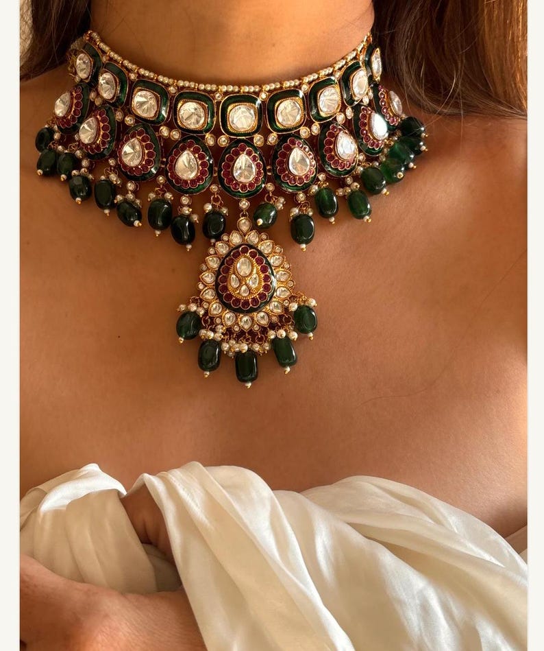 May include: A luxurious choker necklace featuring a gold-toned setting with emerald green and ruby red accents. The necklace is adorned with clear, square-cut stones and teardrop-shaped emerald green beads. A pendant with a similar design hangs from the center.