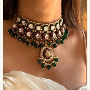 May include: A gold-toned choker necklace with emerald green and clear gemstone accents. The necklace features a central pendant with a teardrop shape and dangling green beads. The design includes red and white accents.