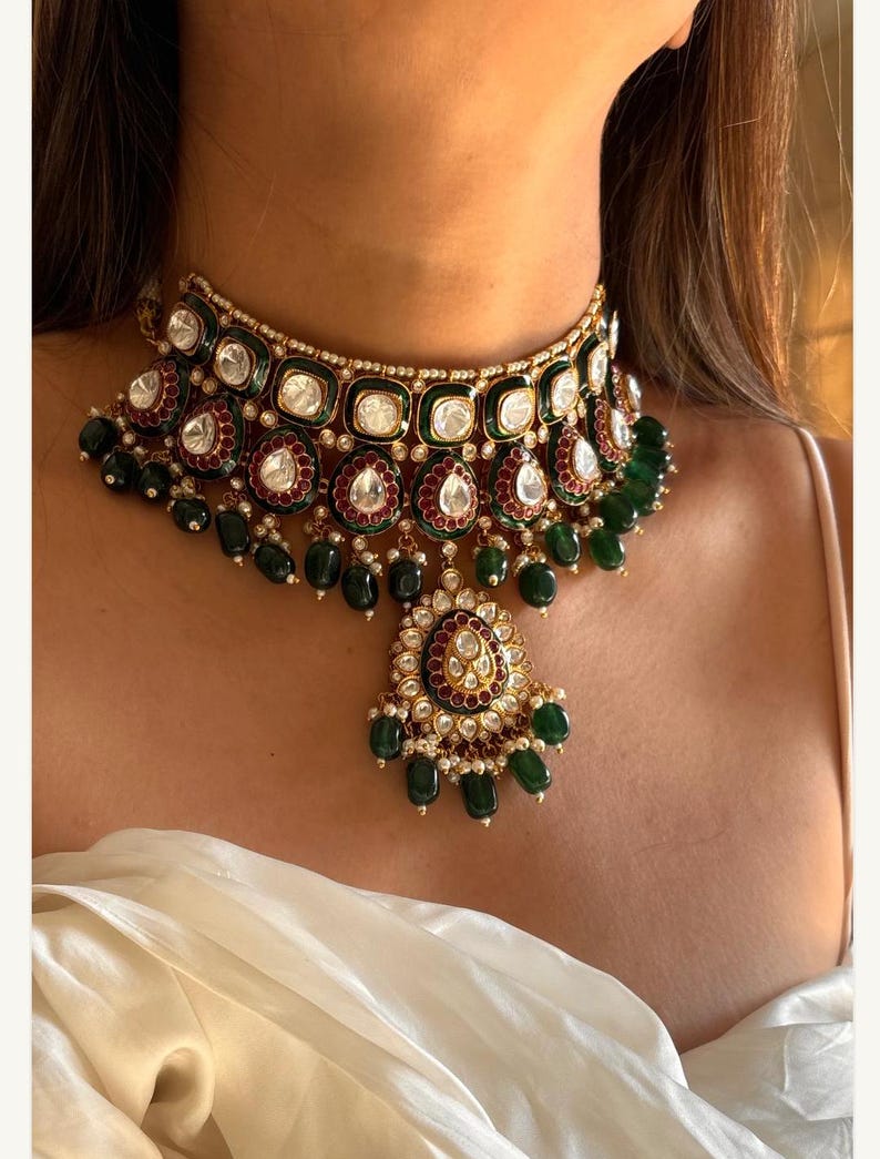 May include: A gold-toned choker necklace with emerald green and clear gemstone accents. The necklace features a central pendant with a teardrop shape and dangling green beads. The design includes red and white accents.