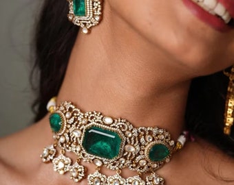 May include: A gold choker necklace with emerald green gemstones and white diamond accents. The necklace has a delicate, intricate design and features a dangling charm.
