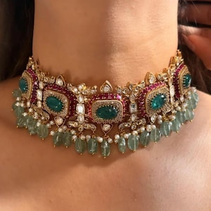 May include: A gold-toned choker necklace featuring emerald green and ruby red gemstones. The necklace has a detailed design with small white gemstones and dangling green beads. The necklace is worn around the neck.