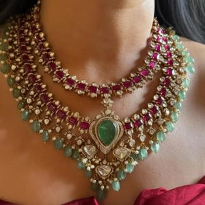 Può includere: Collana multistrato con pietre preziose color rubino e smeraldo. La collana ha una montatura color oro con dettagli intricati. Le pietre preziose sono disposte in modo simmetrico, creando un aspetto lussuoso ed elegante.