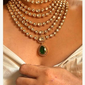 Puede incluir: Un collar de oro de varias capas con un colgante de piedra preciosa verde. El collar está hecho de varias hileras de pequeñas piedras redondas transparentes.