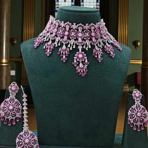 May include: A dazzling jewelry set featuring a necklace and matching earrings. The necklace and earrings are adorned with sparkling white stones and vibrant pink gemstones. The set is displayed on a green velvet bust and stands.
