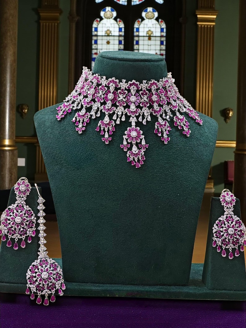 May include: A dazzling jewelry set featuring a necklace and matching earrings. The necklace and earrings are adorned with sparkling white stones and vibrant pink gemstones. The set is displayed on a green velvet bust and stands.
