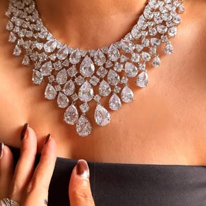 May include: A dazzling diamond necklace featuring a cascading design of various-sized, pear-shaped, and square-cut stones. The necklace is set against a black background, highlighting the brilliance of the clear gemstones. A hand with a ring is visible.