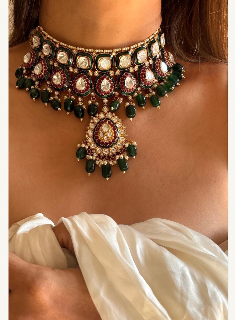 May include: A gold-toned choker necklace with emerald green and ruby red accents. The necklace features square and teardrop-shaped gemstones, with a central pendant. The design is ornate and detailed, with a classic aesthetic.