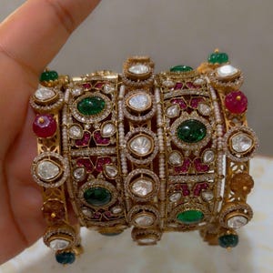 May include: Ornate gold bangle bracelet with intricate detailing. The bracelet features a series of rectangular panels adorned with emerald green and ruby red gemstones, surrounded by clear, sparkling accents. The design incorporates small pearls and additional colorful gems.