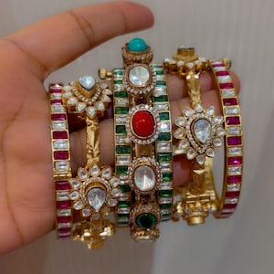 Może przedstawiać: Zestaw czterech złotych bransoletek typu bangle z misternymi zdobieniami metalowymi. Bransoletki są ozdobione różnymi kamieniami szlachetnymi, w tym czerwonymi, zielonymi i turkusowymi kaboszonami oraz przezroczystymi kamieniami o szlifie gruszkowym.
