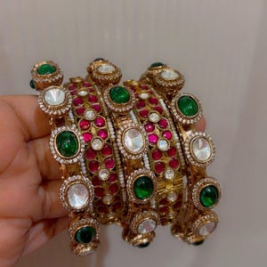 May include: A set of gold-toned bangles featuring alternating oval-shaped green and clear stones. The metal bands are decorated with small, clear pavé-style crystals and rows of small, round, deep pink stones set within the gold-coloured metal frame.