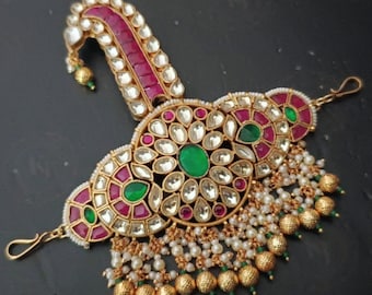 Könnte beinhalten: Ein aufwendiges, goldfarbenes Kopfstück mit einem zentralen floralen Design. Es verfügt über klare, rote und grüne Edelsteine, kleine weiße Perlen und goldene Perlenakzente. Der gebogene obere Teil ist mit roten Steinen besetzt.