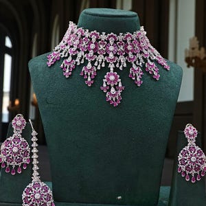 May include: A dazzling jewelry set featuring a necklace, earrings, and a headpiece. The set showcases a floral design with vibrant pink gemstones and sparkling clear stones, all set in a silver-toned metal. The jewelry is displayed on a green velvet bust.