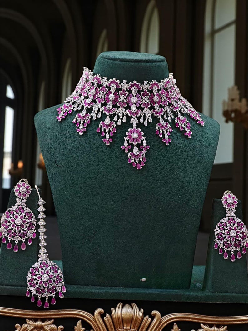 May include: A dazzling jewelry set featuring a necklace, earrings, and a headpiece. The set showcases a floral design with vibrant pink gemstones and sparkling clear stones, all set in a silver-toned metal. The jewelry is displayed on a green velvet bust.