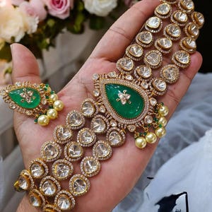 May include: A gold necklace with multiple strands of clear stones and a large green stone pendant. The necklace is on a hand with white pearl beads.