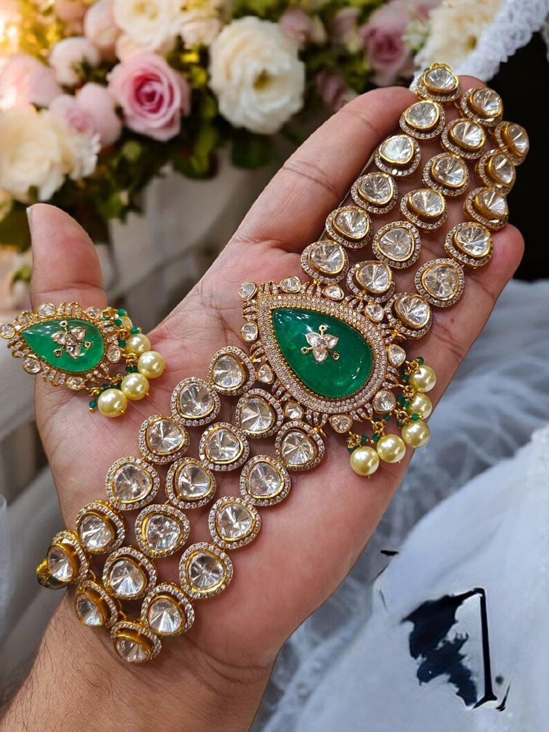 May include: A gold necklace with multiple strands of clear stones and a large green stone pendant. The necklace is on a hand with white pearl beads.