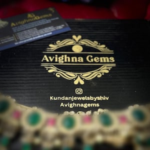 May include: A black box with the gold text "Avighna Gems" and a business card on top. The box has a textured surface. A necklace with green and pink stones is in the foreground.