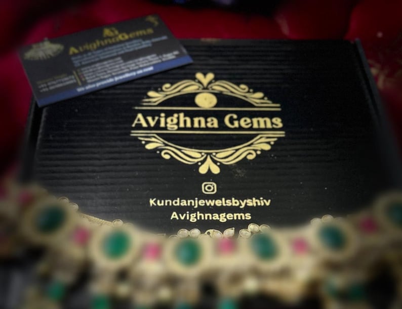 May include: A black box with the gold text "Avighna Gems" and a business card on top. The box has a textured surface. A necklace with green and pink stones is in the foreground.