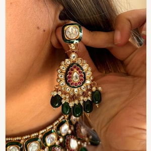 May include: Gold-toned statement earrings with emerald green and ruby red accents. The earrings feature a large, ornate design with dangling green beads and small white pearls. The top of the earring has a square emerald green border with a clear stone.