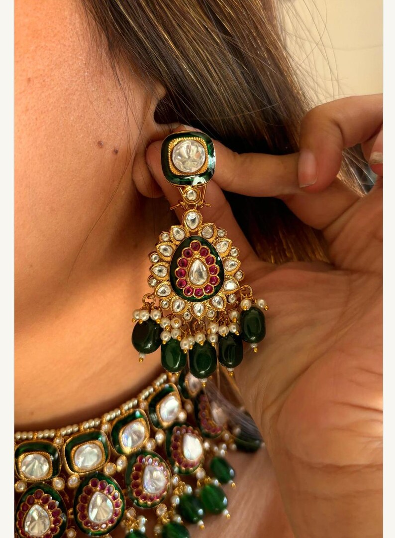 May include: Gold-toned statement earrings with emerald green and ruby red accents. The earrings feature a large, ornate design with dangling green beads and small white pearls. The top of the earring has a square emerald green border with a clear stone.