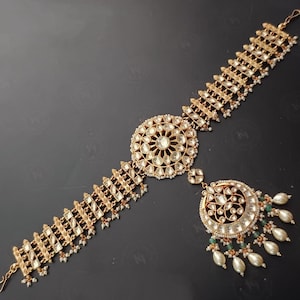 May include: A gold-toned headpiece with a large, round, jeweled centerpiece and a dangling pendant with a smaller, round, jeweled design. The headpiece is adorned with white pearls and green beads.