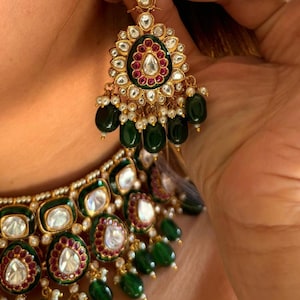 May include: Close-up of a gold-toned jewelry set featuring an earring and necklace. The earring has a floral design with a central white stone, surrounded by red and green accents, and dangling green beads. The necklace mirrors the earring's design.