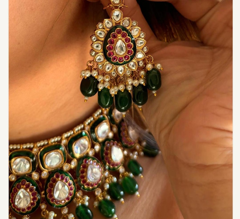 May include: Close-up of a gold-toned jewelry set featuring an earring and necklace. The earring has a floral design with a central white stone, surrounded by red and green accents, and dangling green beads. The necklace mirrors the earring's design.