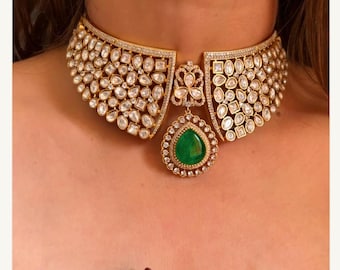 Op de afbeelding: Een goudkleurige choker ketting met een centrale smaragdgroene hanger. De ketting is versierd met heldere, sprankelende stenen in verschillende vormen. De hanger heeft een druppelvorm, omringd door kleinere stenen. Een opvallend sieraad.