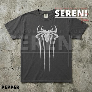 Puede incluir: Camiseta gris oscuro con un dise&ntilde;o de ara&ntilde;a blanca. El gr&aacute;fico es una ara&ntilde;a estilizada con l&iacute;neas que gotean. La camiseta tiene cuello redondo y mangas cortas. La palabra "SERENI" es visible en la esquina superior derecha.