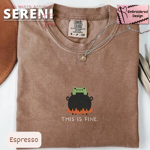 May include: Espresso-colored t-shirt with an embroidered design of a frog on a cauldron with the text "THIS IS FINE." The shirt has a Comfort Colors tag and a silver necklace. The design is on the front of the shirt.