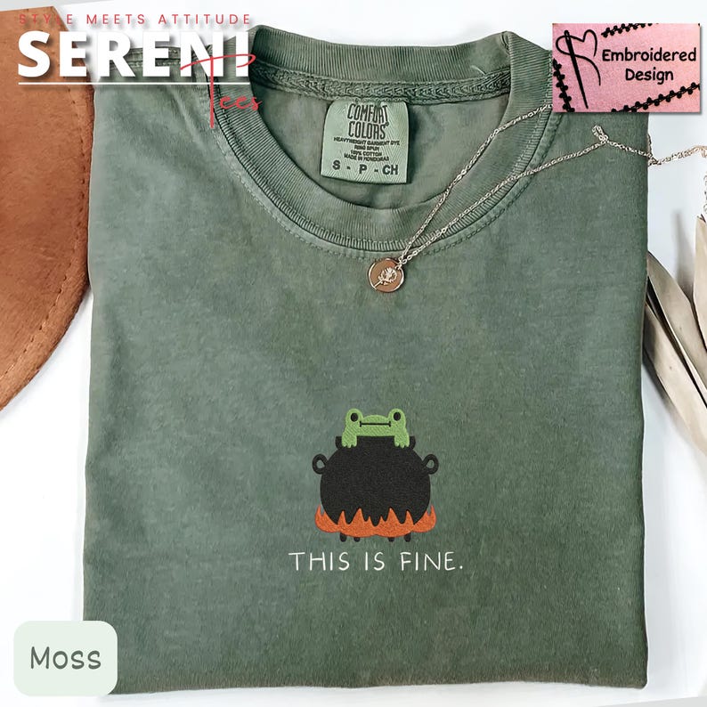 May include: Moss green t-shirt with a frog and cauldron graphic, and the text "THIS IS FINE." The shirt has a crew neck and short sleeves. A silver necklace with a small pendant is on the shirt. The shirt is labeled "Embroidered Design."