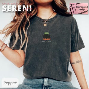 May include: Dark grey t-shirt with a small embroidered design of a frog in a cauldron above the text "THIS IS FINE." The cauldron has orange flames at the bottom. The shirt is a comfortable, casual style.