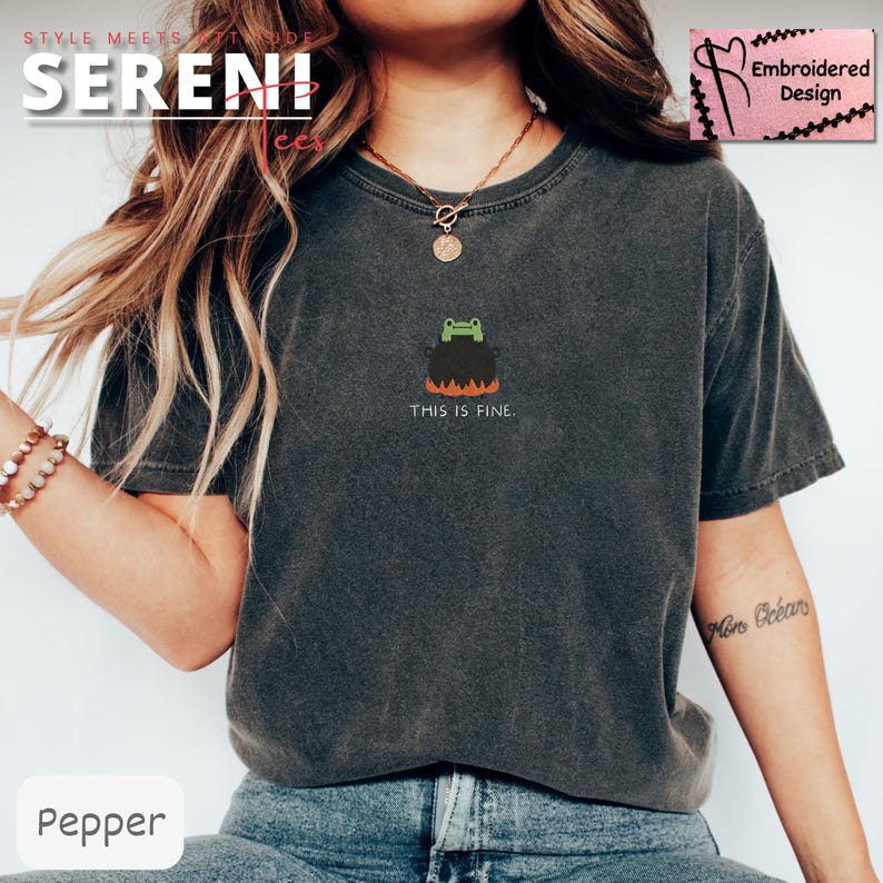 May include: Dark grey t-shirt with a small embroidered design of a frog in a cauldron above the text "THIS IS FINE." The cauldron has orange flames at the bottom. The shirt is a comfortable, casual style.