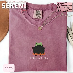 May include: Berry-colored t-shirt with a crew neck. The shirt features an embroidered design of a frog on a cauldron with the text "THIS IS FINE." A silver necklace with a pendant is also visible.